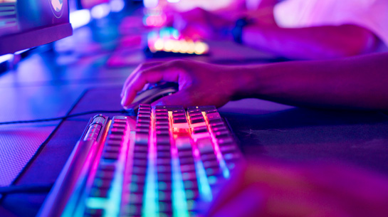 A designer building brands and websites in a dark room with purple-pink lights, typing on a lighted rainbow keyboard and using a mouse