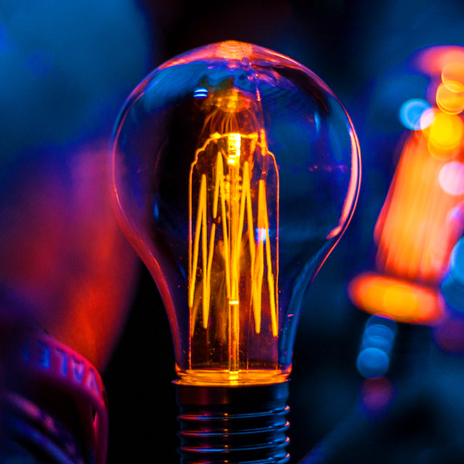 A light bulb glowing orange and reflecting pink on a navy blue background representing creativity and design thinking AI cannot replace