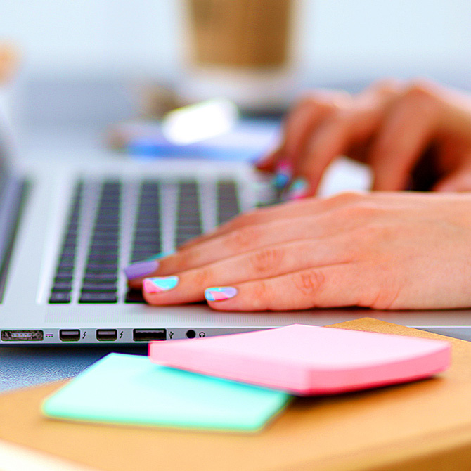 A woman's hands with brightly painted nails using a laptop to define the scope of work for a project, with post-it notes and a coffee cup nearby