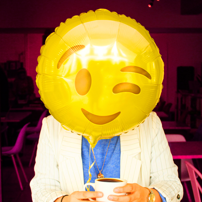 A confident website owner wxcited to sharfe her website represented by a woman in a blue shirt and while blazer holding a coffee mug with a bright yellow winky-face emoji, mylar helium balloon in front of her face
