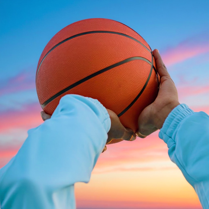 A man's arms in a blue sweatshirt about to shoot a basketball with a colorful sunset in the background