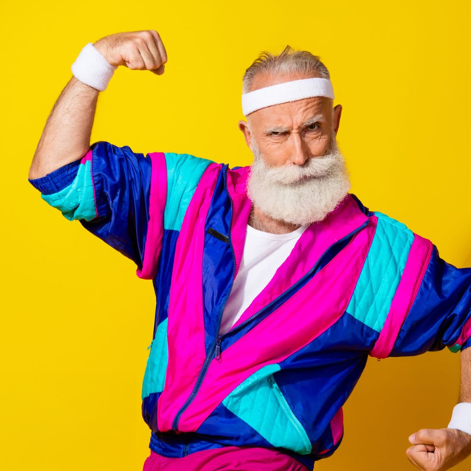 A senior-aged man with a white mustache and beard in a blue, turquoise, and pink 80s track suite and sweat band, flexing his muscles