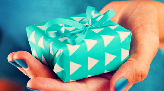 A woman in a teal sweater and dark teal painted fingernails holding a package wrapped in in light, bright teal wrapping paper with white triangles and teal curly ribbon