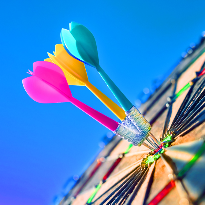 Bright pink, yellow, and turquoise darts all in the center bullseye — the one core focus — of a dartboard