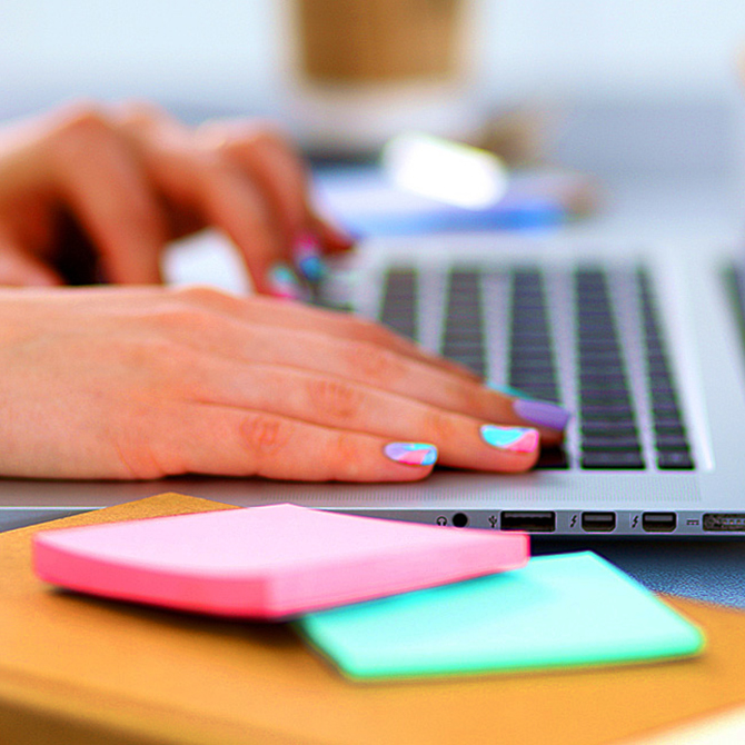 A woman with brightly painted fingers nails designing marketing materials for a client on a laptop with pink and teal sticky notes sitting on top of a notebook