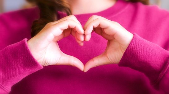 A brunette woman in a pink sweatshirt making a heart with her hands to show that she loves small projects